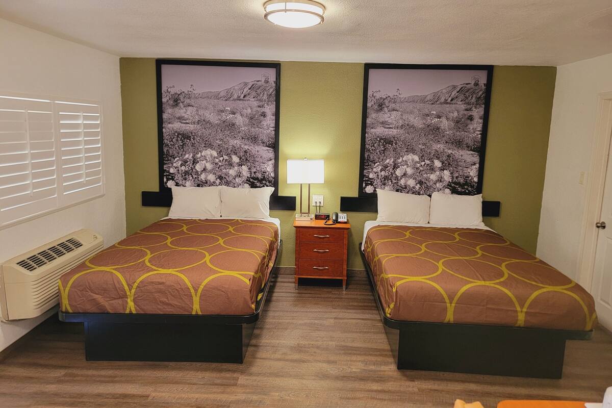 A hotel room with two beds, a nightstand, a lamp, and a large window with blinds.
