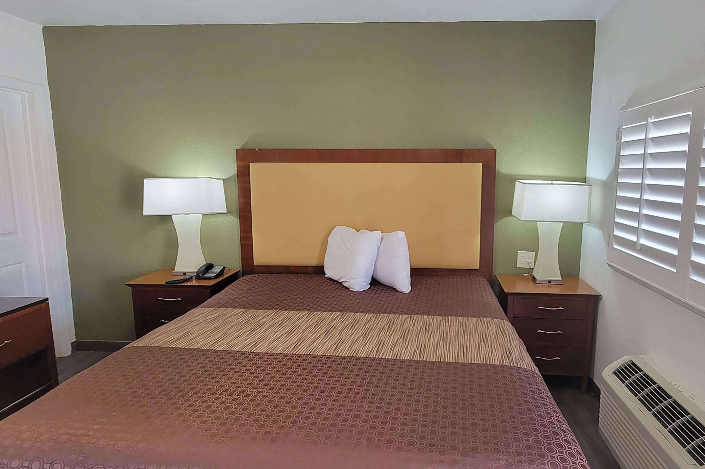 A hotel room with a bed, two bedside tables with lamps, a nightstand, and a window with blinds.