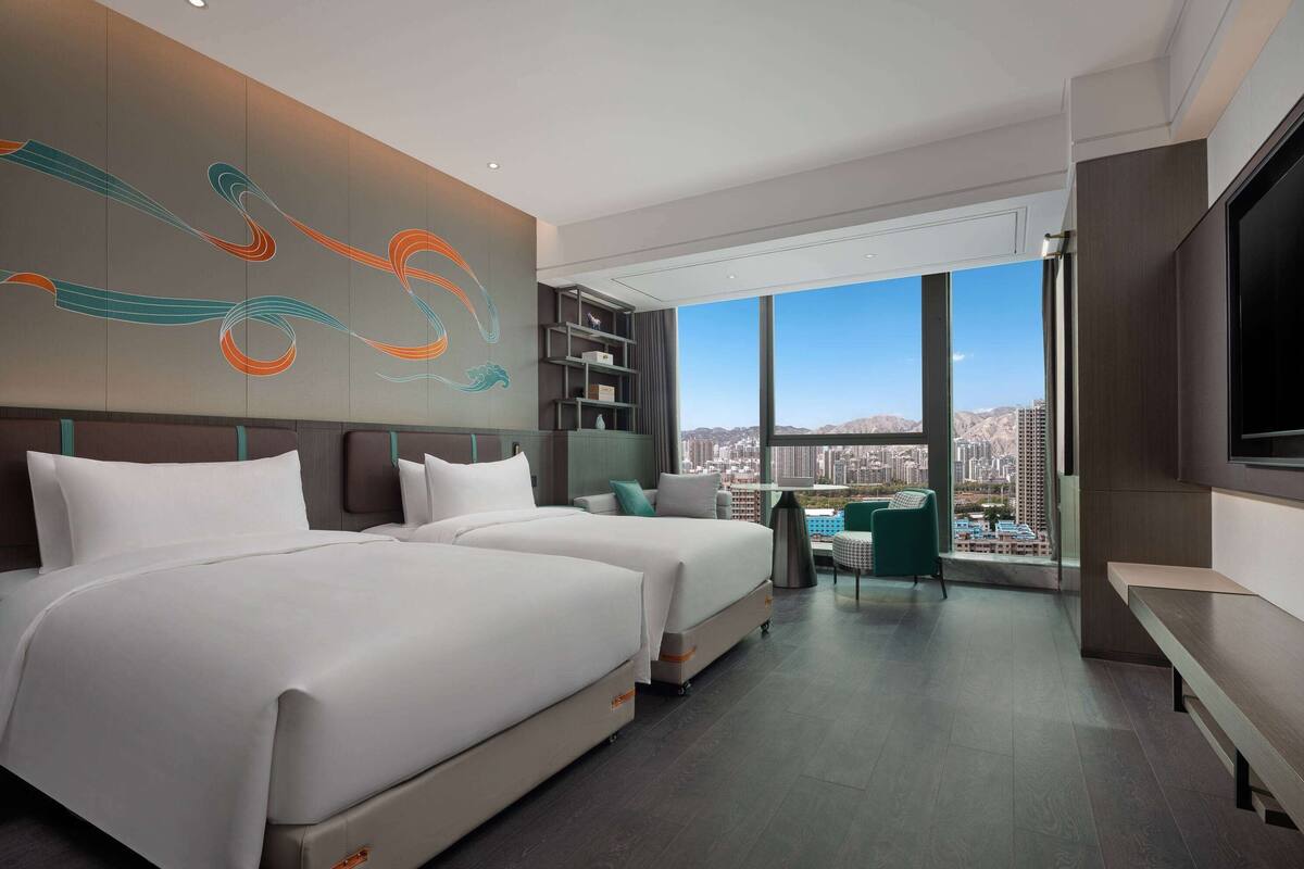 A modern hotel room with two beds, a desk, and a large window offering a city view.
