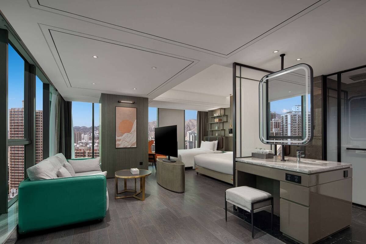 A modern hotel room with a large window, a sofa, a bed, a TV, a desk, and a bathroom with a mirror.