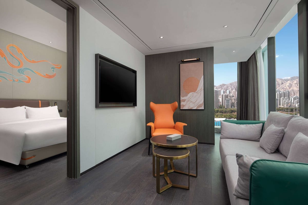 A modern hotel room with a bed, a sofa, an orange armchair, a small round table, and a large TV.