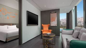 A modern hotel room with a bed, a sofa, an orange armchair, a small round table, and a large TV.