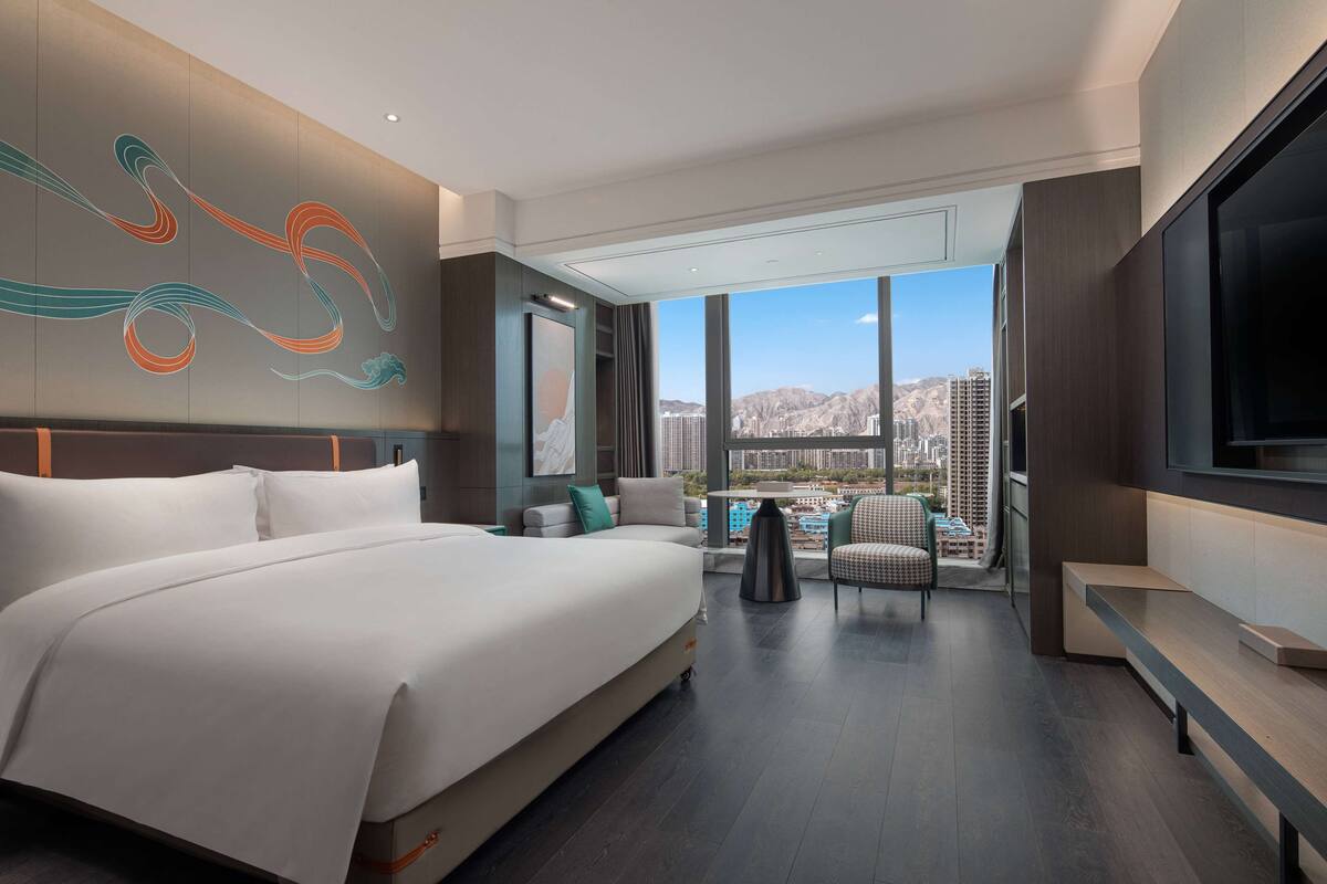A modern hotel room with a large bed, a TV, and a view of the cityscape.