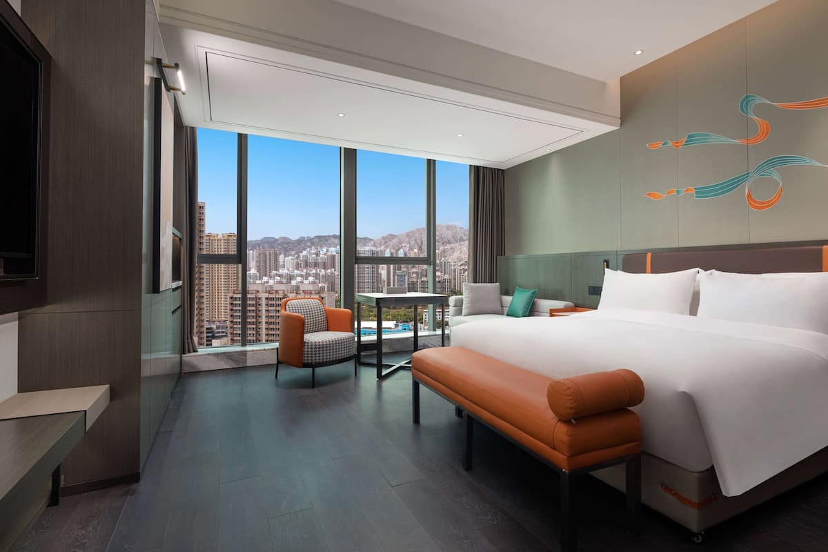 A modern hotel room with a large bed, a seating area, and a balcony with a city view.