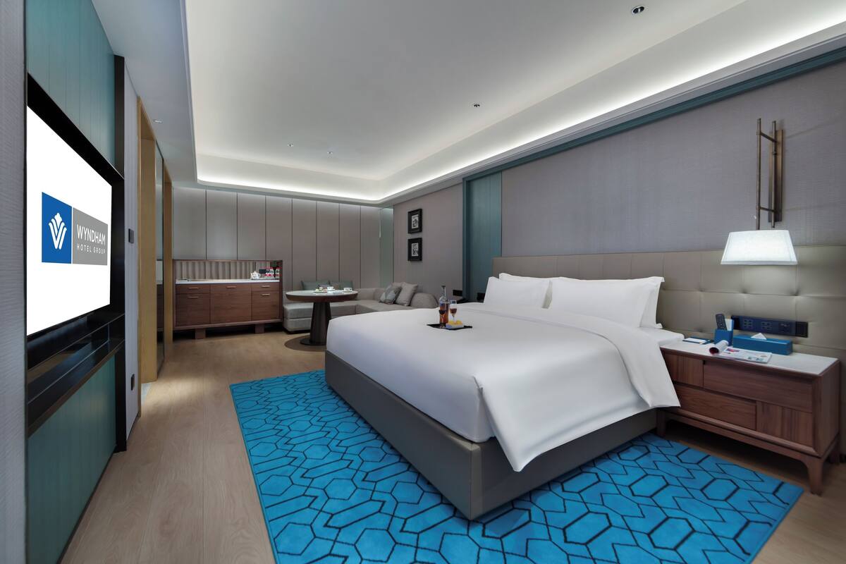 Una habitación de hotel moderna con una cama grande, una mesita de noche con lámpara, un pequeño escritorio y una zona de estar con sofá y mesa de centro.