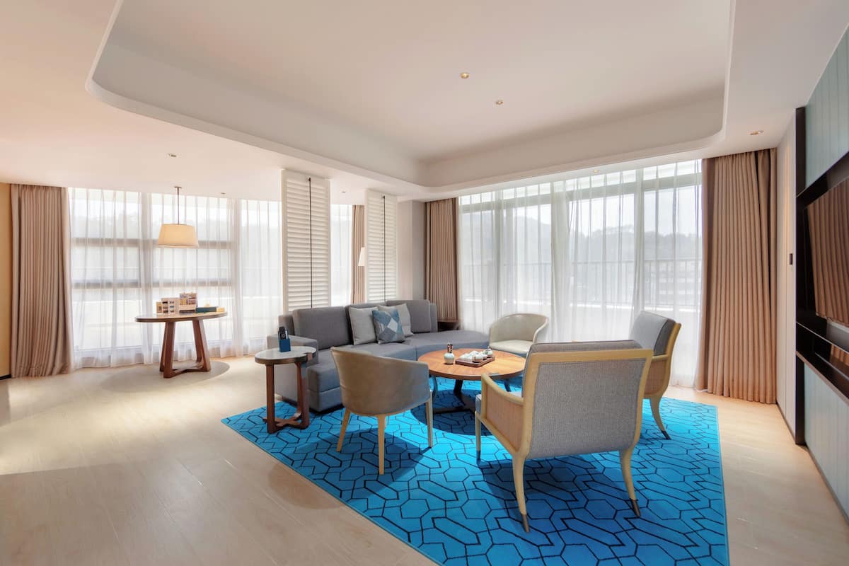 Habitación de hotel moderna con sofá, dos sillones, una mesa de centro y una mesita auxiliar. Amplias ventanas con cortinas dejan entrar luz natural.