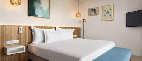 A modern hotel room with a large bed, wooden headboard, bedside tables, a wall-mounted TV, and artwork.