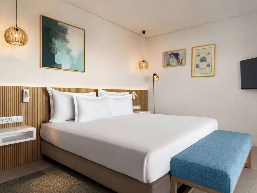 Una habitación de hotel moderna con una cama grande, cabecero de madera, mesitas de noche, televisor montado en la pared y una obra de arte.