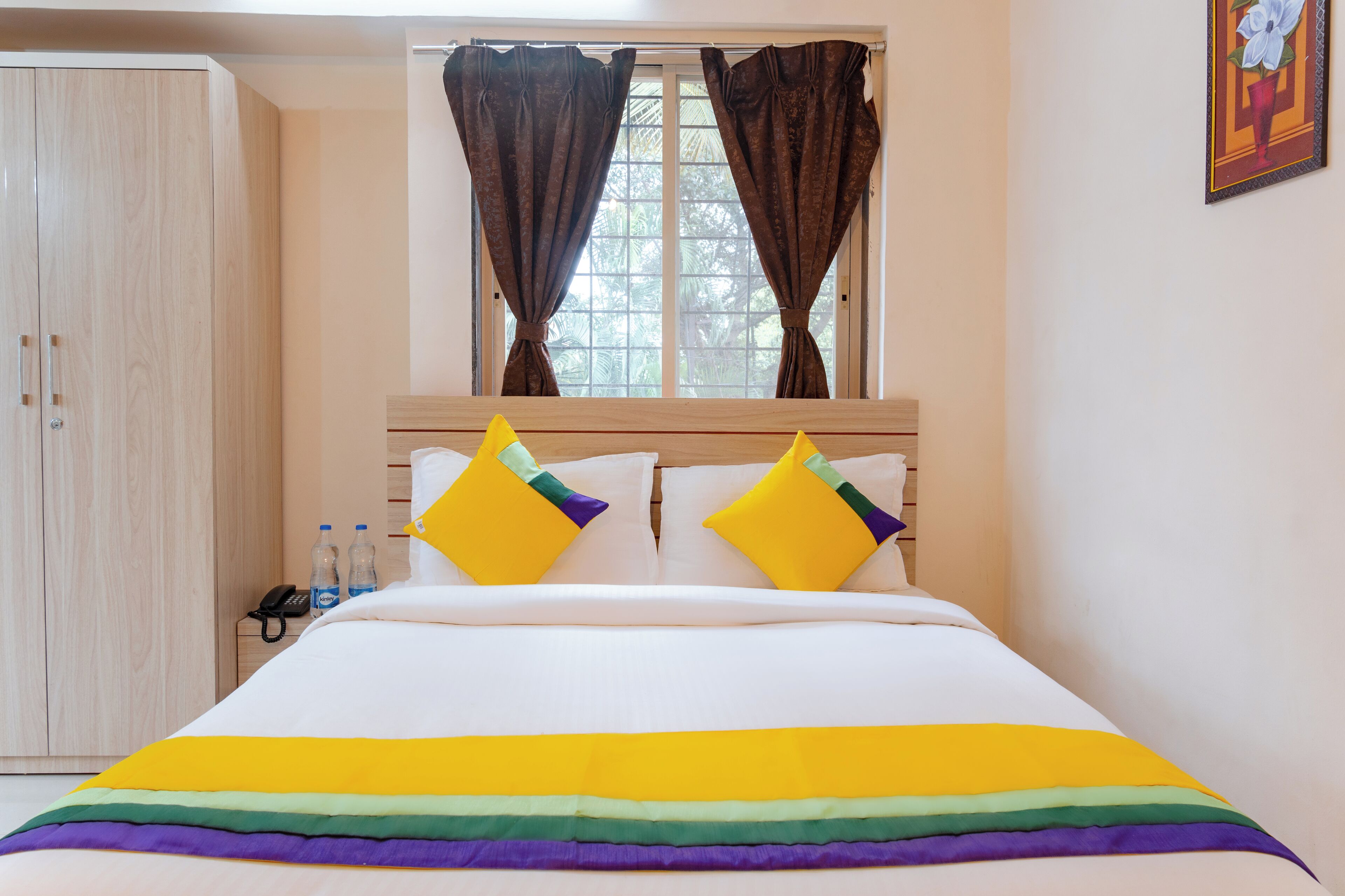 A hotel room with a bed, a wardrobe, a window with curtains, and a painting on the wall.