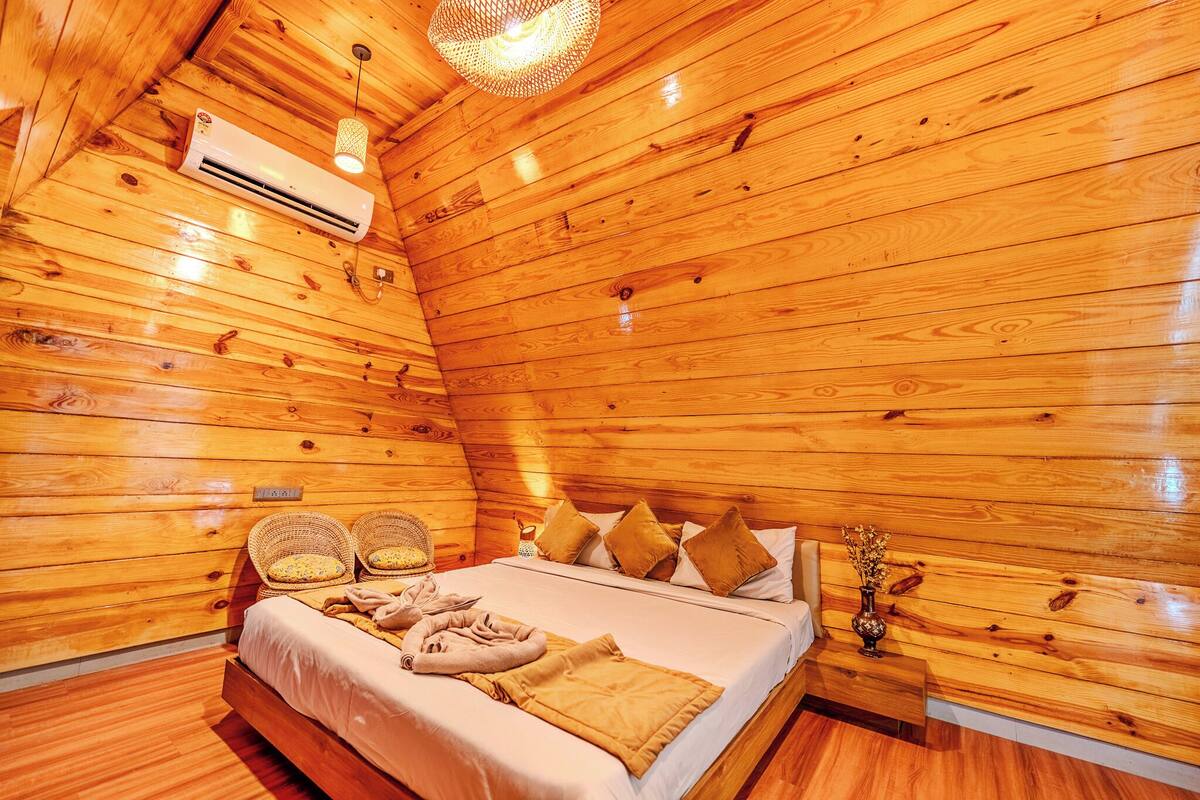 Une pièce de style cabane en bois comprenant un lit, deux fauteuils en osier, une table de chevet avec un vase et un appareil de climatisation.