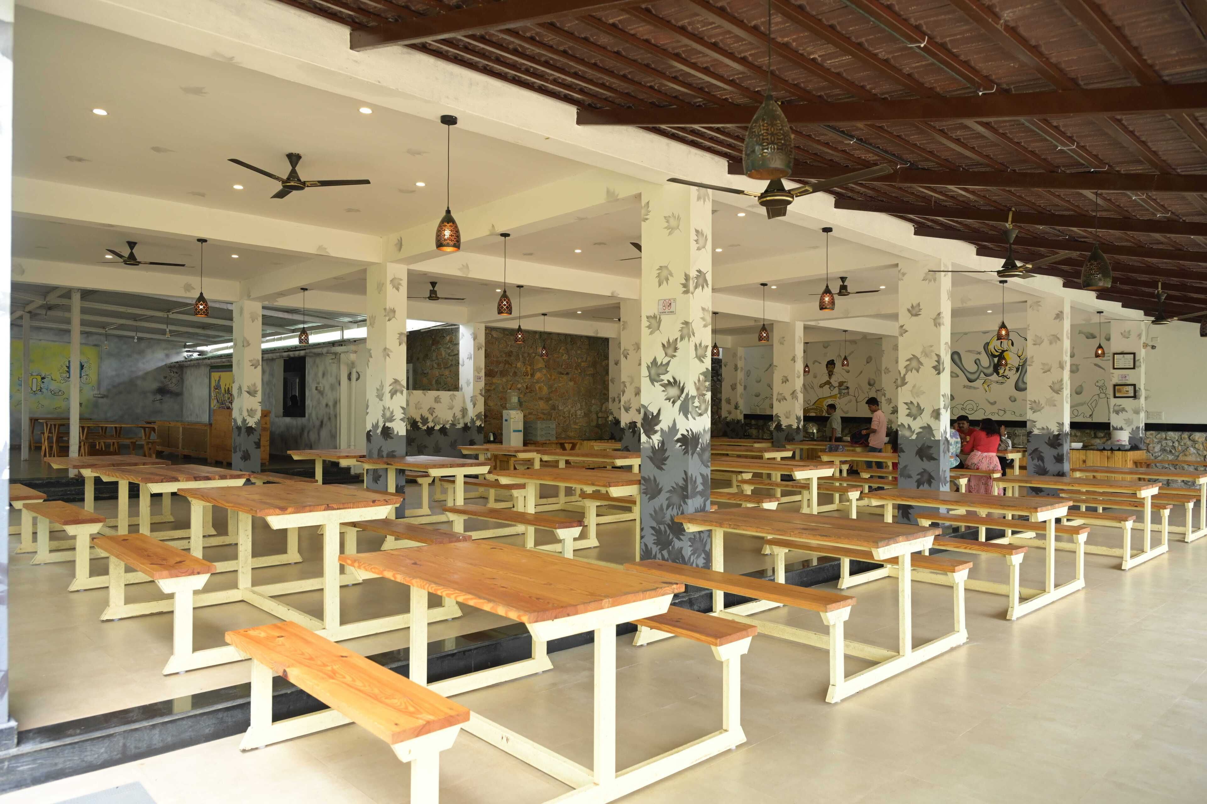 A spacious hall with rows of wooden benches and tables, ceiling fans, and decorative hanging lights.