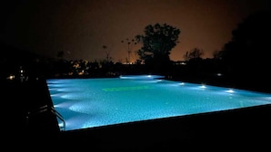 A nighttime view of a swimming pool with illuminated edges, surrounded by trees and distant lights.