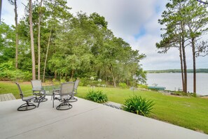 Pátio com mesa e cadeiras, vista para um lago, rodeado por árvores e vegetação.
