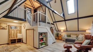 Een ruime loft met hoge plafonds, zichtbare balken, een houten trap, een gezellige zithoek met een leren fauteuil en een klein boekenrek.