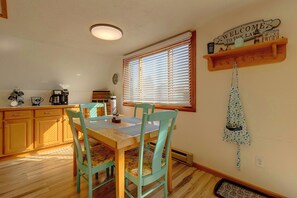 Een eetruimte met een houten tafel en stoelen, een raam met zonwering, een klok en een welkomstbord aan de muur.