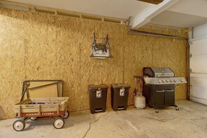 A garage with a wooden wall, a metal cart, two trash bins, and a barbecue grill.