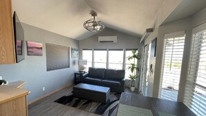 A modern living room with a sofa, ottoman, and a dining area with a wooden table. There are large windows and a ceiling fan. - Elephant Lake Sunset Oasis (Elephant Butte)