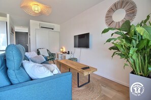 A modern living room with a blue sofa, a wooden coffee table, a large potted plant, a TV mounted on the wall, and a round mirror. - Nouveaute Appartement Neuf "thinic", VUE MER à 100m de la Plage (Saint-Pierre-Quiberon)