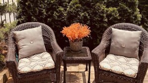 Two wicker chairs with cushions, a small table with a vase of orange flowers, and a backdrop of tall green trees.