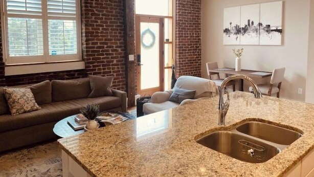 A modern living room with a brown leather sofa, a coffee table, and a kitchen area with granite countertops and a stainless steel sink.