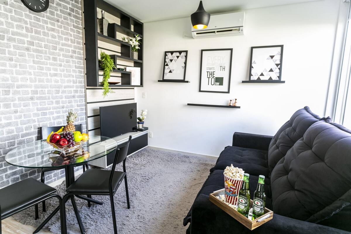 A modern living room with a glass table, black chairs, a black sofa, and a wooden tray with bottles and popcorn.