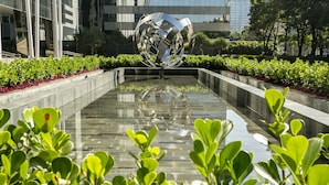 Una escultura moderna en una plaza con superficie acuática reflectante, rodeada de vegetación y edificios al fondo.