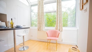 A modern kitchen with white cabinets, a wooden countertop, a pink chair, and a window with curtains. - Cute&Cozy Barons Courth Studio Apartment (London)