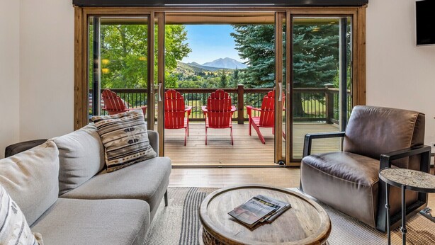 Un salon comprenant un canapé, un fauteuil et une table basse. Une porte-fenêtre coulissante donne sur une terrasse extérieure avec des chaises rouges et une vue sur les arbres et les montagnes.