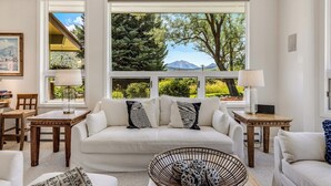 Un salon avec un canapé blanc, deux tables d’appoint avec des lampes, un fauteuil en osier et une vue sur les arbres et les montagnes, visible à travers de grandes fenêtres.