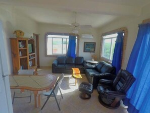 Relax in this inviting living room with a sofa, table, chairs, and bookshelf, surrounded by large windows and a beautiful painting.