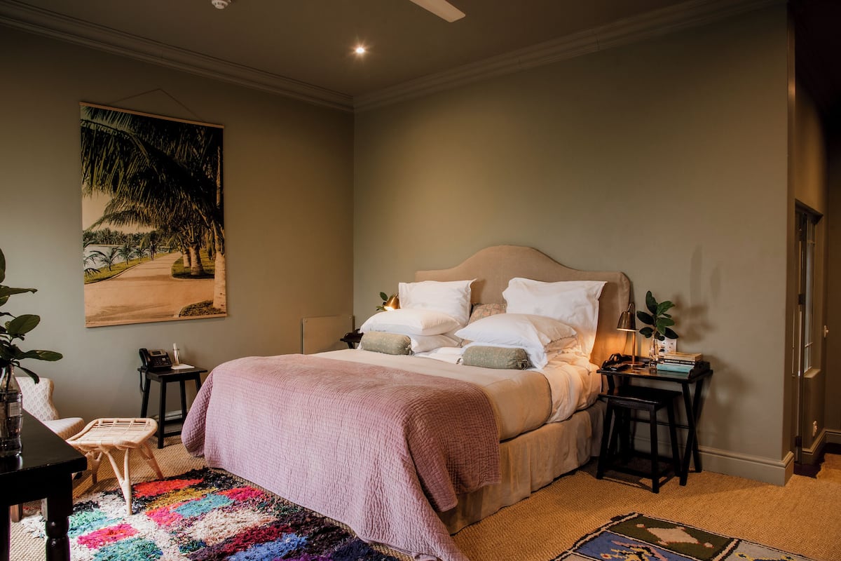A bedroom with a large bed, a bedside table with a lamp, a small chair, and a colorful rug.