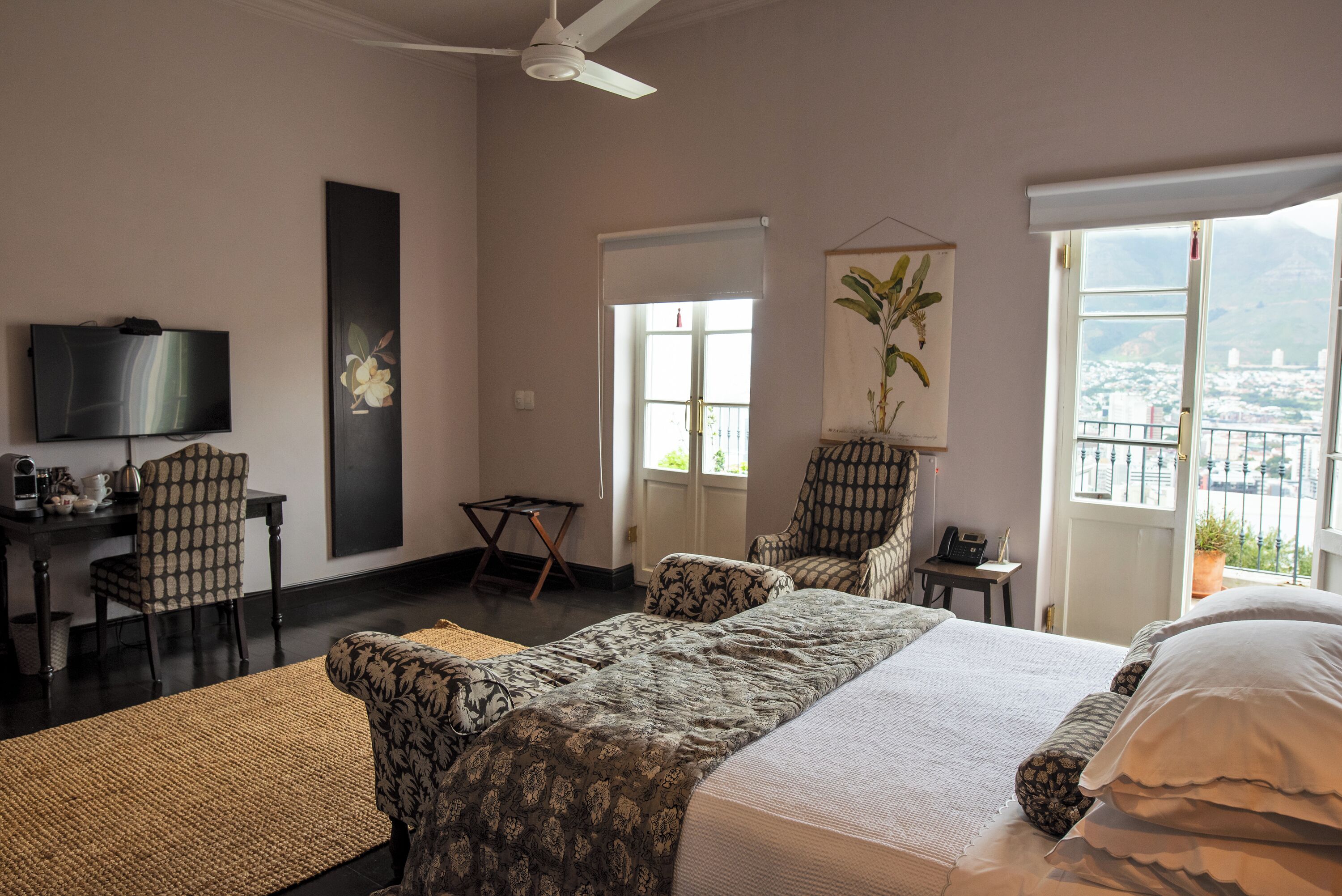 A hotel room with a bed, two chairs, a desk with a television, a window with a view, and a ceiling fan.