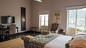 A hotel room with a bed, two chairs, a desk with a television, a window with a view, and a ceiling fan.