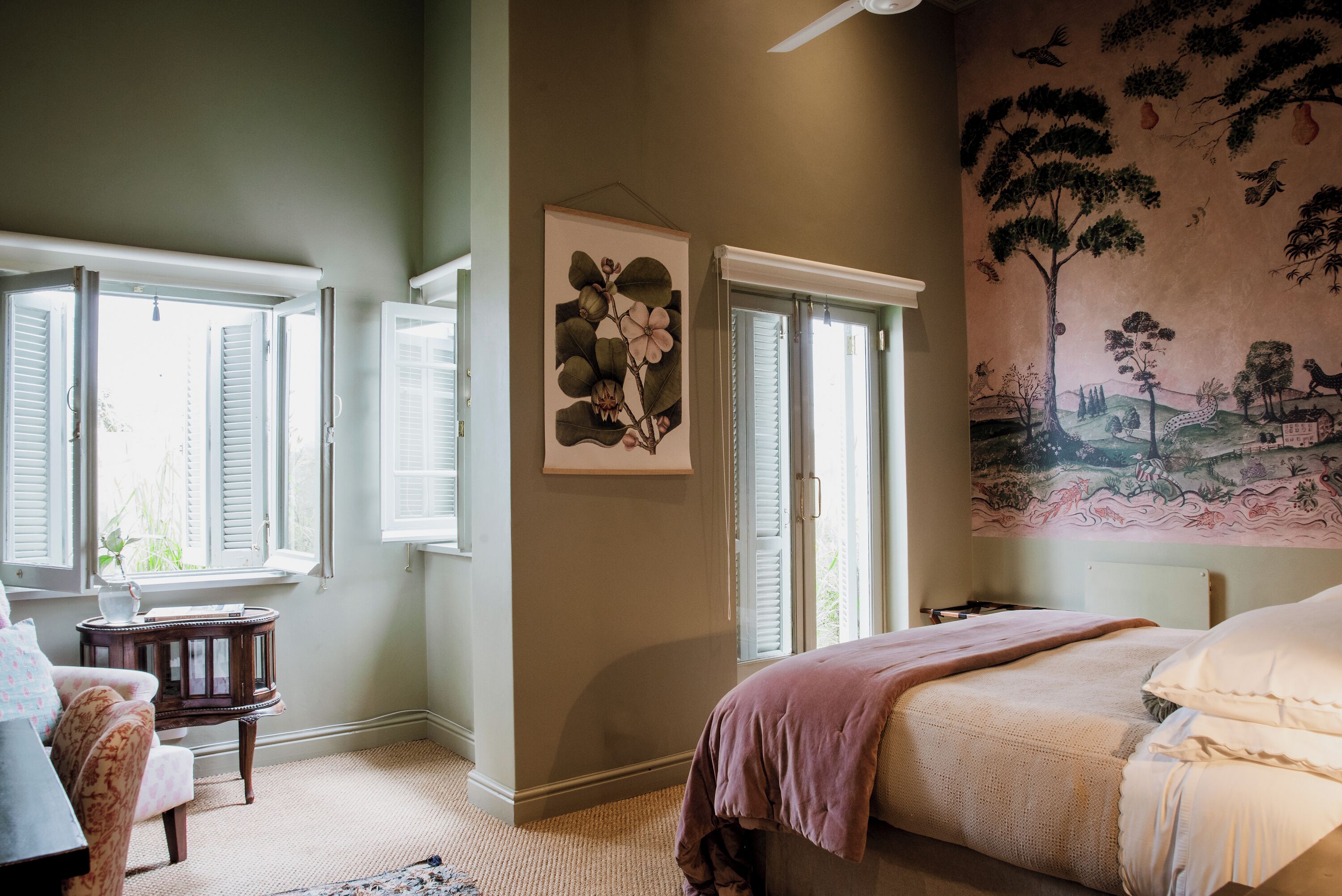 A bedroom with a bed, a window with shutters, a wall-mounted artwork, and a small table with a lamp.