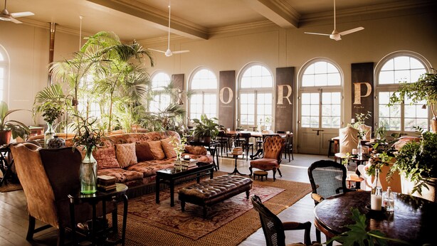 A spacious lounge with a large sofa, armchairs, and a coffee table, surrounded by potted plants and natural light from arched windows. - Dorp Hotel (Cape Town)