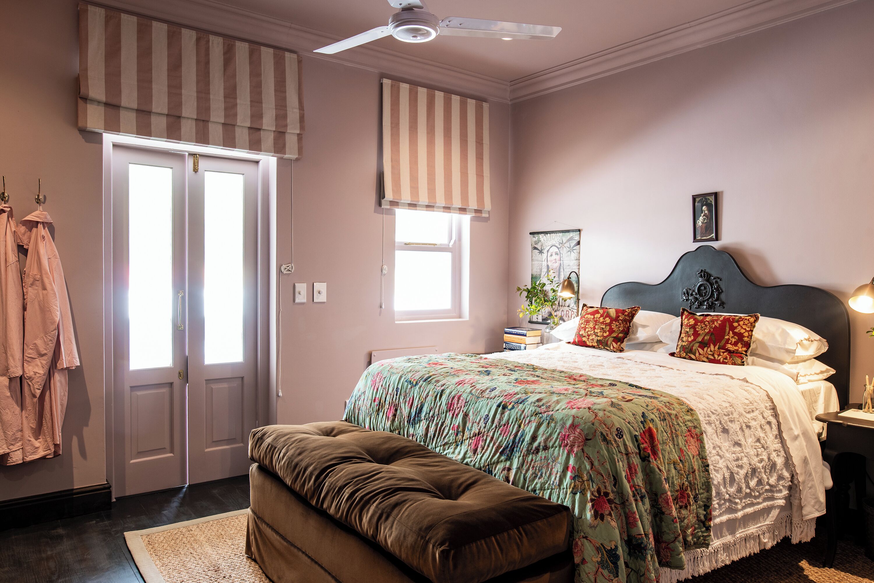 A bedroom with a bed, a bench, a ceiling fan, a window with blinds, and a door.