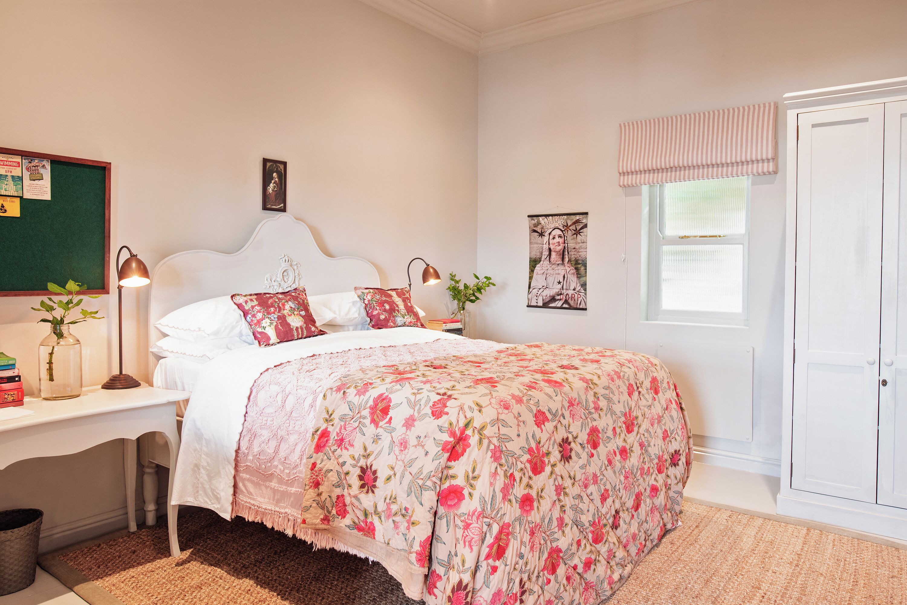 A bedroom with a bed, a desk with a lamp, a plant, and a window with a curtain.