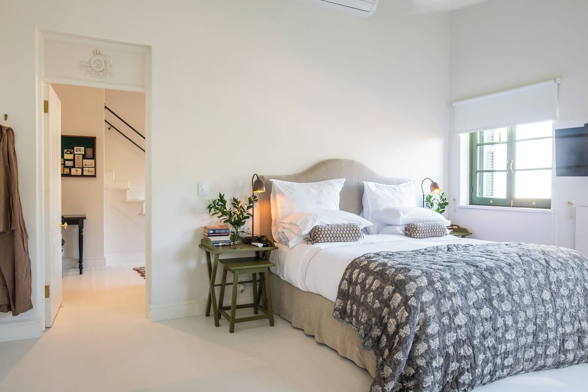 A bedroom with a bed, bedside tables, a nightstand, a window with blinds, and a staircase leading to another room.