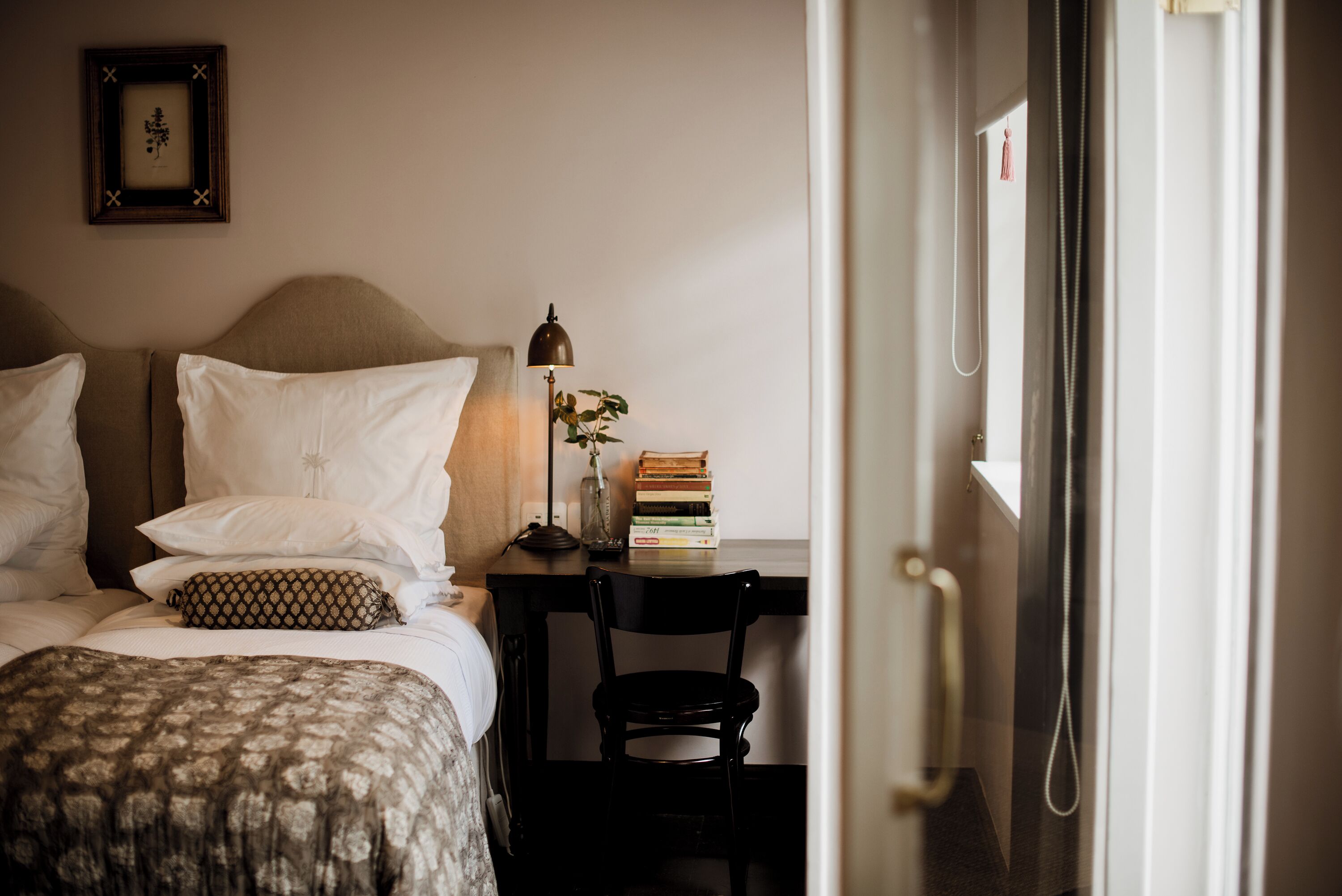 A neatly made bed with pillows, a bedside lamp, a stack of books, and a chair.
