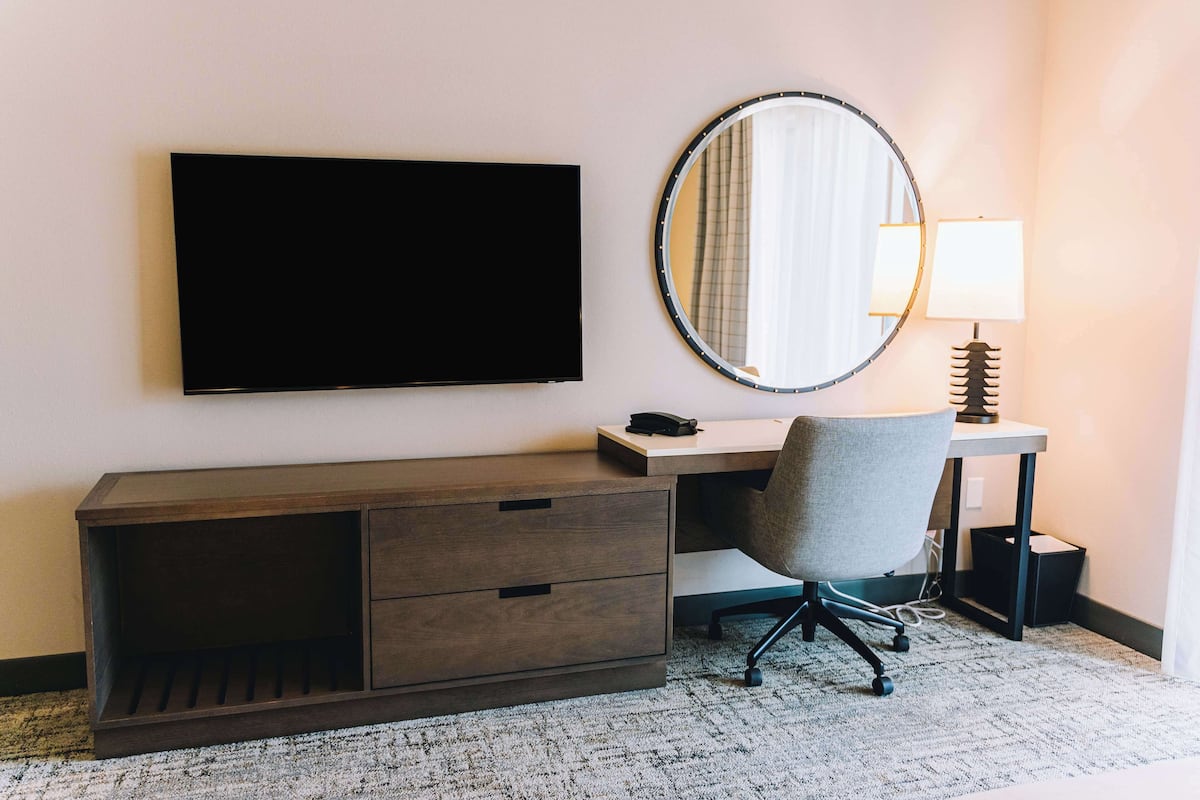 A hotel room with a flat-screen TV mounted on the wall, a desk with a chair, a round mirror, and a lamp.