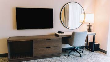 A hotel room with a flat-screen TV mounted on the wall, a desk with a chair, a round mirror, and a lamp.