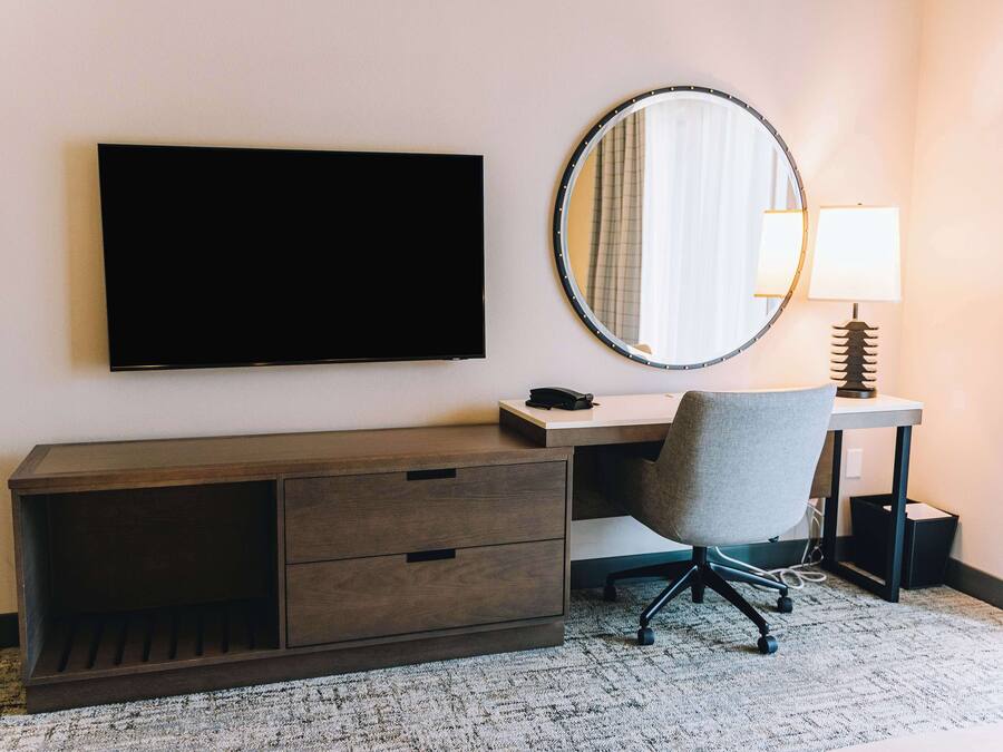 A hotel room with a flat-screen TV mounted on the wall, a desk with a chair, a round mirror, and a lamp.