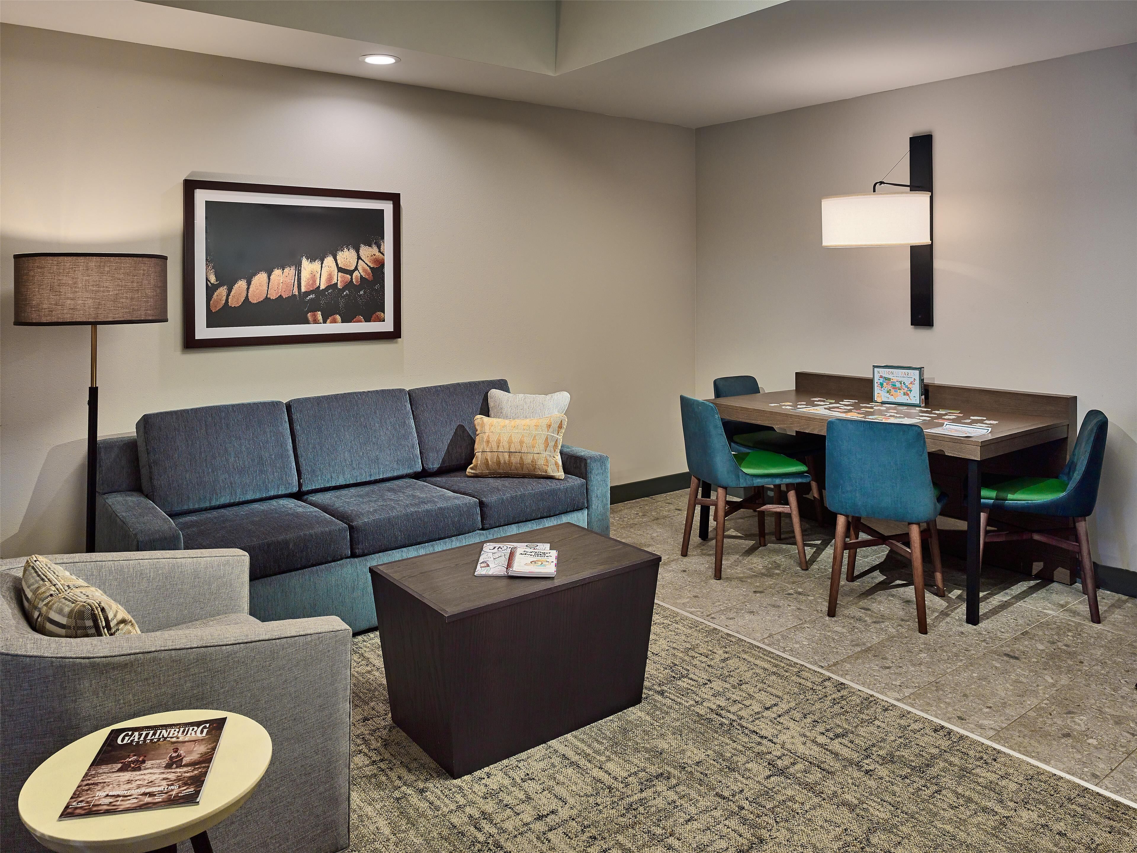 A modern hotel room with a gray sofa, a small round table with a magazine, a dark coffee table, a dining area with blue chairs and a wooden table, and a framed artwork on the wall.