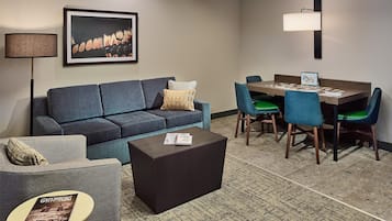 Ein modernes Hotelzimmer mit grauem Sofa, einem kleinen runden Tisch mit einer Zeitschrift, einem dunklen Couchtisch, einem Essbereich mit blauen Stühlen und einem Holztisch sowie einem gerahmten Kunstwerk an der Wand.