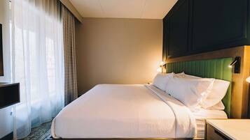 A hotel room with a neatly made bed, a bedside table, a television, and a window with sheer curtains.