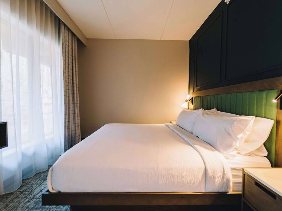 A hotel room with a neatly made bed, a bedside table, a television, and a window with sheer curtains.