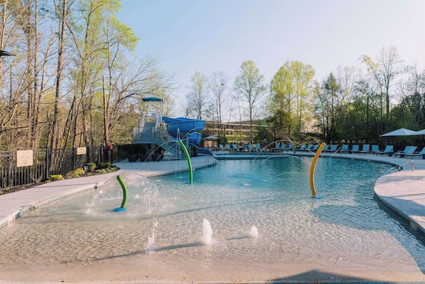 Una piscina con tobogán de agua, rodeada de árboles y sillas de descanso.