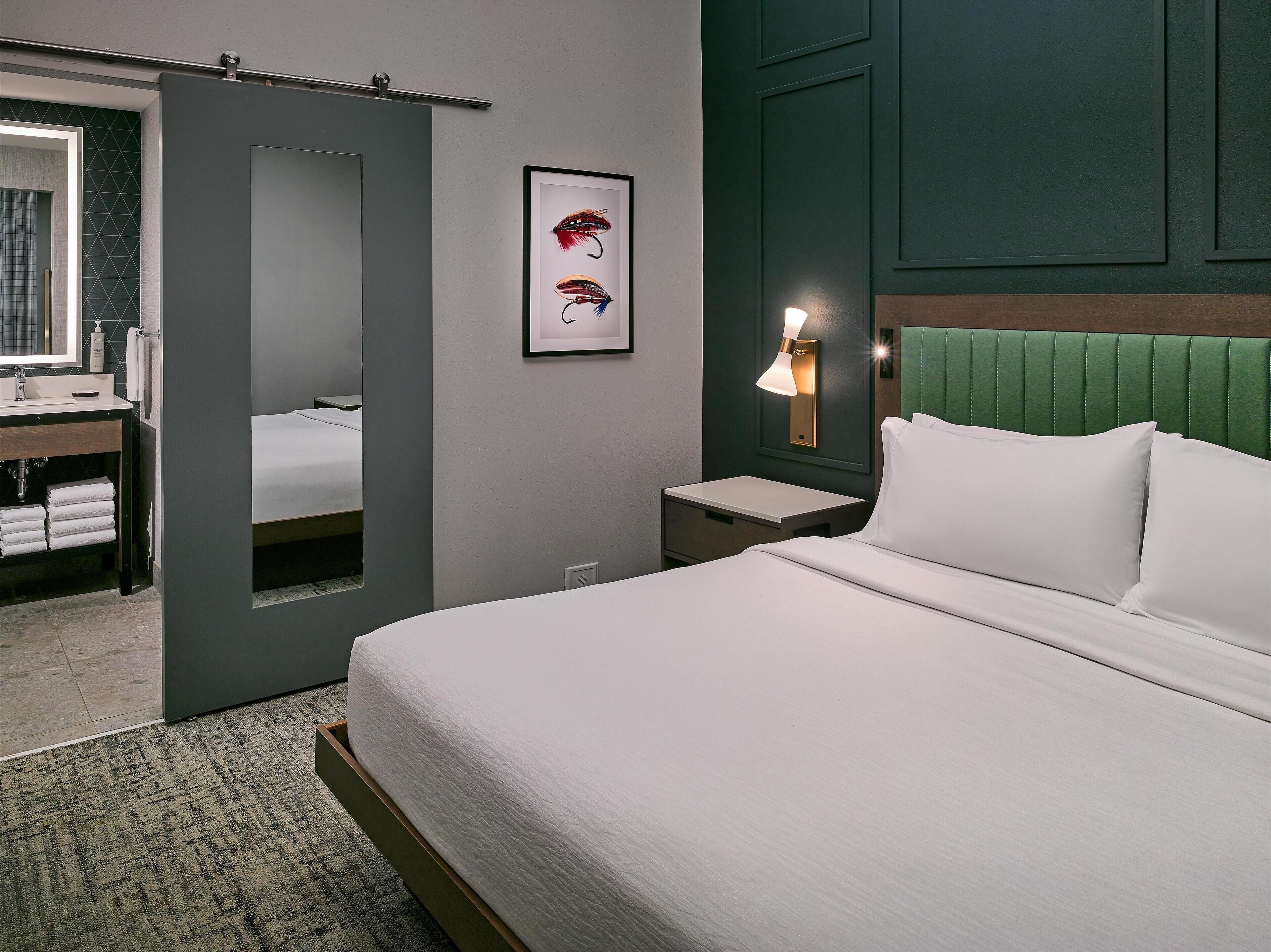 Habitación de hotel con cama, mesita de noche, lámpara de pared, cuadro con señuelos de pesca, baño con espejo y toallero.