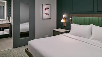 A hotel room with a bed, a nightstand, a wall-mounted lamp, a framed picture of fishing lures, a bathroom with a mirror, and a towel rack.