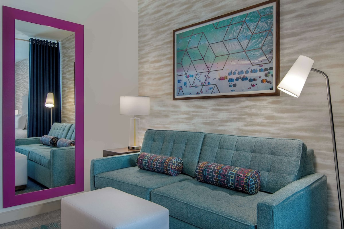A teal sofa with patterned cushions, a large mirror with a purple frame, a geometric artwork on the wall, and a floor lamp.